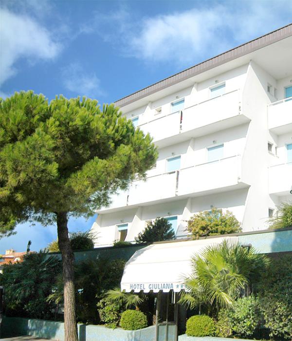 Hotel moderno con balconi e giardino verde, cielo azzurro sopra.